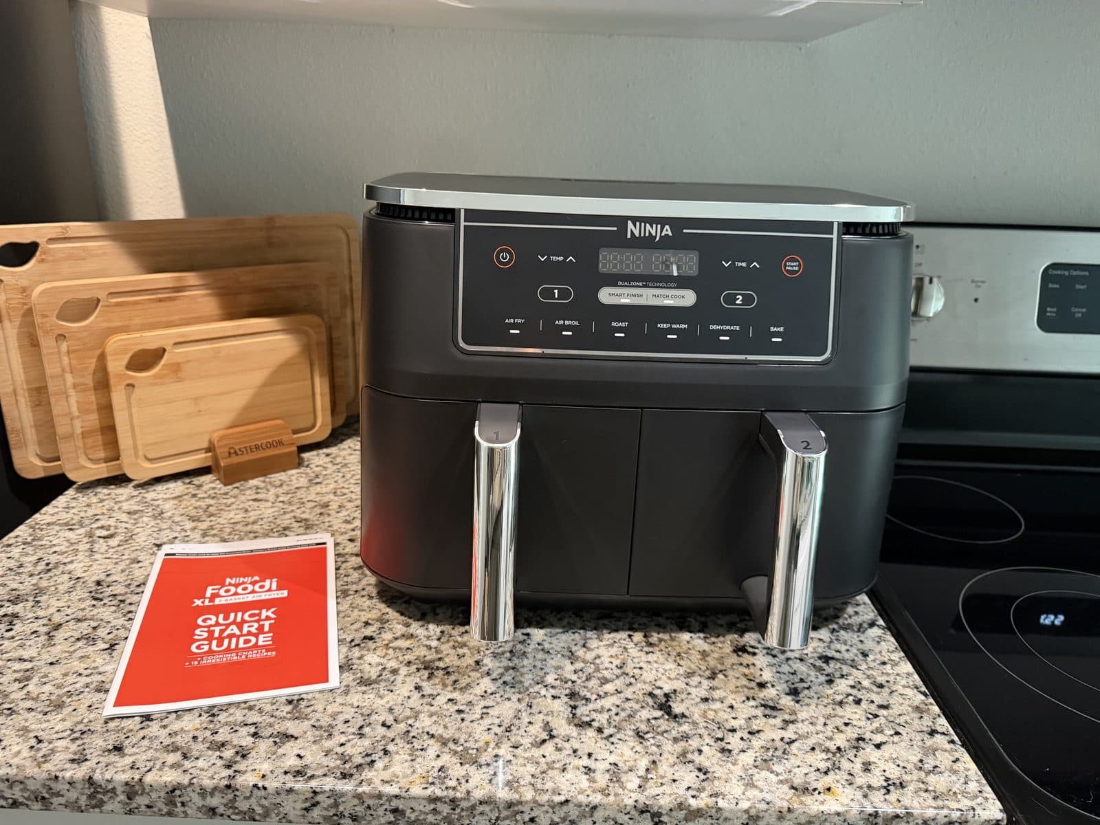 A black Ninja Foodi XL DualZone air fryer with two front baskets and brushed-steel handles on a granite kitchen counter, beside wooden cutting boards and a red quick start guide.