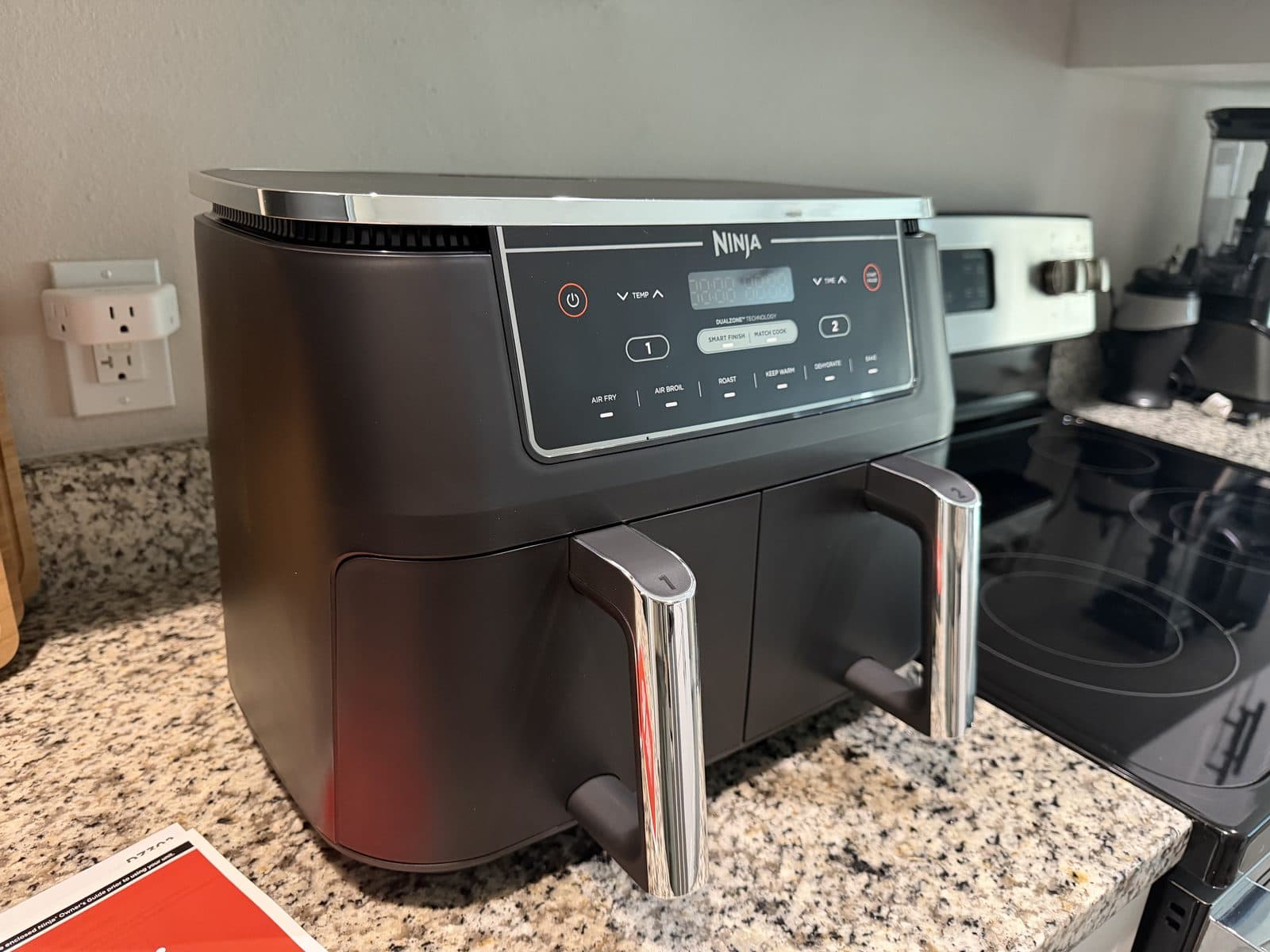 A three-quarter view of the Ninja Foodi XL DualZone air fryer on a granite counter, showing its black body, steel-trimmed lid and two basket handles.