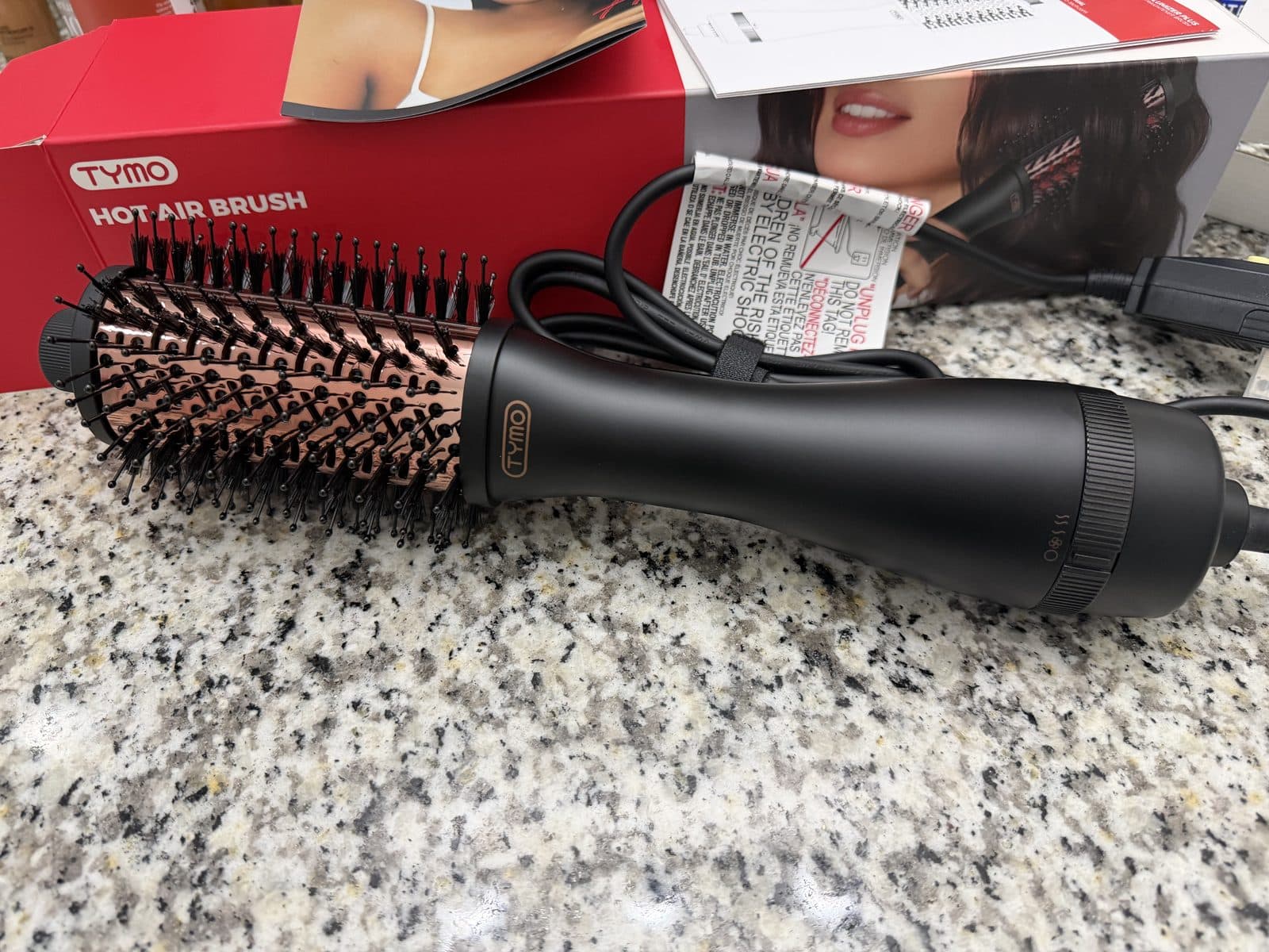 A black TYMO Volumizer Plus hot air brush with a rose-gold oval barrel and bristles, lying on a speckled granite counter in front of its red TYMO box.