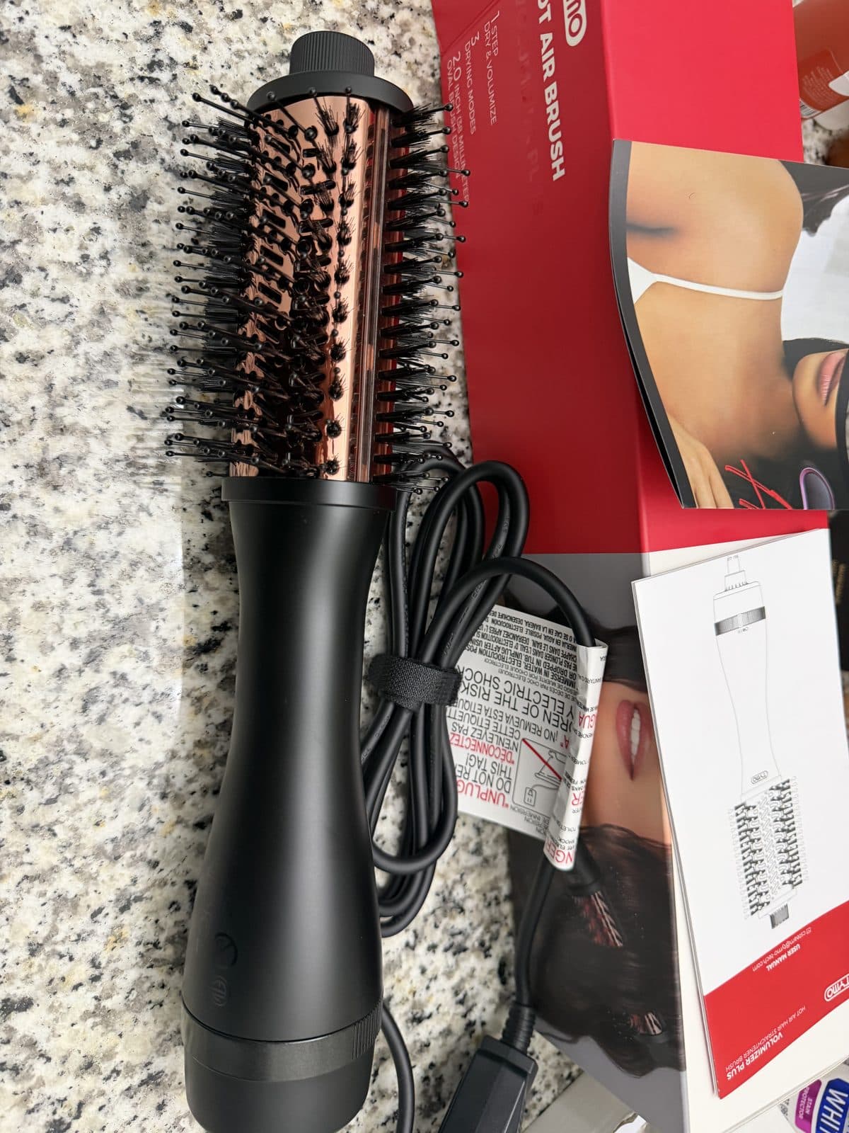 Close-up of the TYMO Volumizer Plus oval brush head, showing rose-gold barrel and black bristles, resting on a granite counter beside its box.