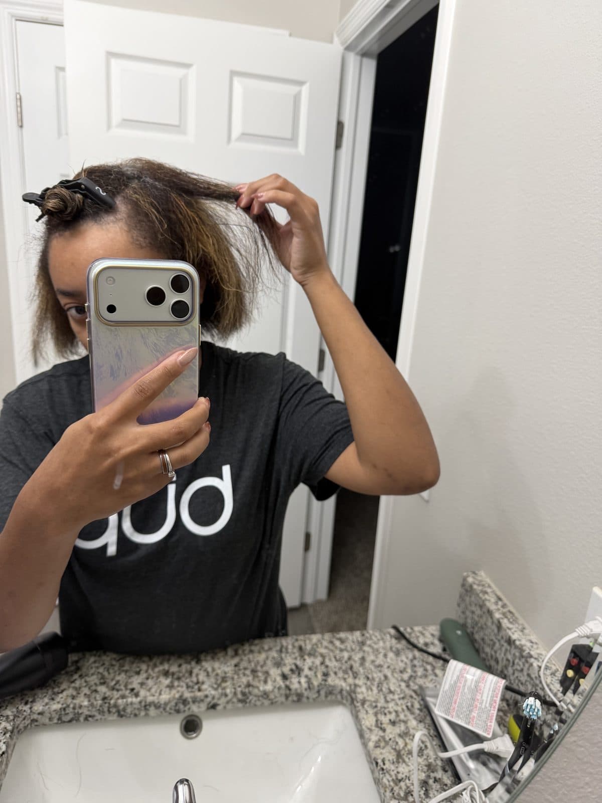 Mirror selfie of Martika sectioning her chin-length hair at a granite bathroom vanity, the top layer clipped up and one section lifted between her fingers.